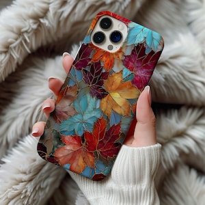 May include: A colorful phone case featuring a stained-glass design of autumn leaves in shades of red, orange, and blue. The case has a glossy finish and a gold outline around each leaf. The phone case is held in a person's hand.