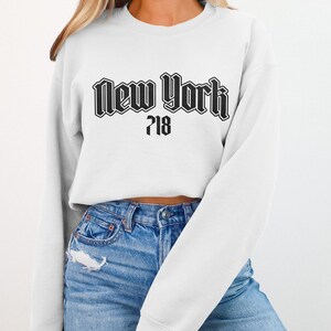May include: White sweatshirt with the text "New York 718" in a black gothic font. The sweatshirt is paired with blue jeans. The image focuses on the upper body of the person wearing the sweatshirt.
