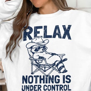 May include: A white crewneck sweatshirt featuring a dark blue graphic of a raccoon wearing a cowboy hat and sunglasses while lounging in a folding chair. The text above reads RELAX and the text below reads NOTHING IS UNDER CONTROL.