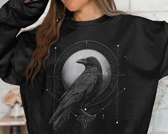Peut inclure: Un sweat-shirt noir à col rond arborant une impression graphique d'un corbeau centré dans un cadre circulaire. Le motif comprend des lignes géométriques et de petits points entourant l'oiseau, représenté de profil sur un fond circulaire gris clair.