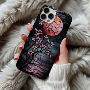 May include: A black phone case with a stained glass design featuring a pink cherry blossom tree and a red and orange moon.