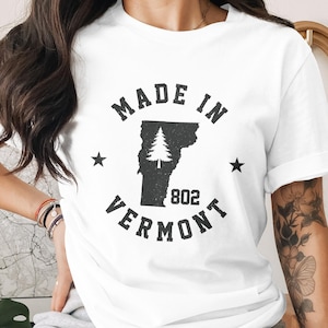 May include: White t-shirt with a black graphic featuring the text "MADE IN VERMONT" in a semi-circle, with the state outline and a tree graphic in the center. The number "802" is below the tree.