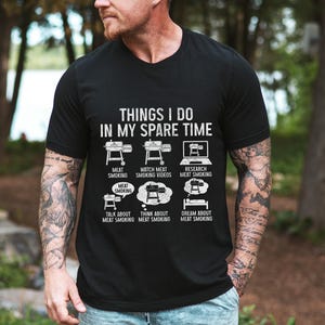 May include: Black t-shirt with white text and graphics about meat smoking. The text reads "THINGS I DO IN MY SPARE TIME" with illustrations of smokers, a laptop, and thought bubbles. The shirt's theme is barbecue and grilling.