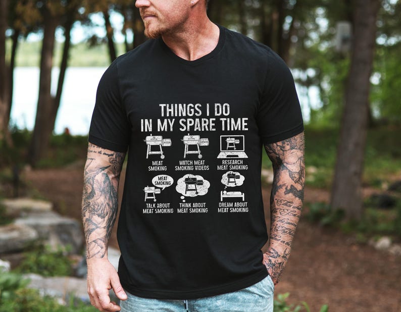 May include: Black t-shirt with white text and graphics about meat smoking. The text reads "THINGS I DO IN MY SPARE TIME" with illustrations of smokers, a laptop, and thought bubbles. The shirt's theme is barbecue and grilling.