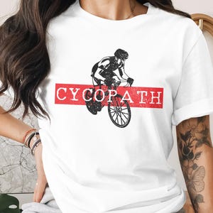 Può includere: T-shirt bianca con grafica di ciclista in nero e rosso. La parola "CYCOPATH" è stampata in bianco su uno striscione rosso. Il ciclista è raffigurato in bicicletta.