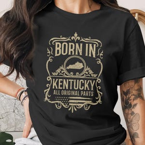 Może przedstawiać: Czarny t-shirt z wzorem w stylu vintage. Wzór zawiera napis "BORN IN KENTUCKY" nad konturem stanu Kentucky, z napisem "ALL ORIGINAL PARTS" poniżej. Projekt jest w kolorze złotym.