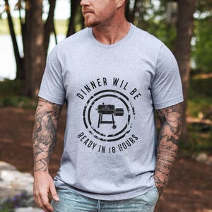 K&ouml;nnte beinhalten: Ein graues T-Shirt mit einer schwarzen Grafik eines Grills und dem Text "Dinner will be ready in 18 hours".