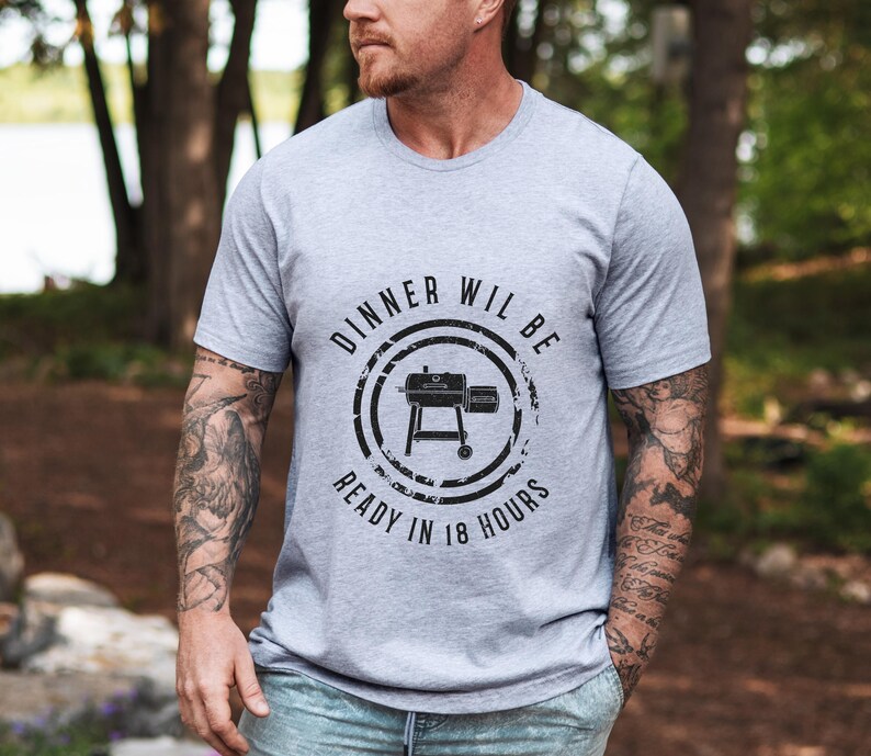 K&ouml;nnte beinhalten: Ein graues T-Shirt mit einer schwarzen Grafik eines Grills und dem Text "Dinner will be ready in 18 hours".
