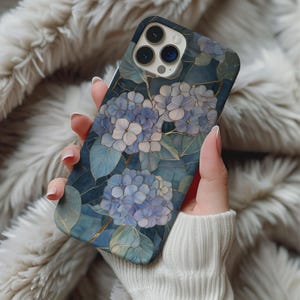 May include: A smartphone case featuring a floral design with blue and white hydrangea blooms and green leaves. The case has a dark blue background with gold accents. The phone case is held in a person's hand.