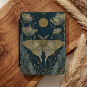 Può includere: Una copertina di taccuino blu scuro con un'illustrazione di falena dorata ed elementi floreali. Il design include una luna dorata e fiori stilizzati. La copertina ha un dorso nero ed è ambientata su uno sfondo di legno.