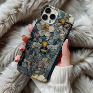 May include: A phone case featuring a floral and bee design. The case has a dark teal background with a pattern of flowers in shades of white, pink, and yellow. Two bees are visible, with yellow and black stripes. The case is held in a person's hand.