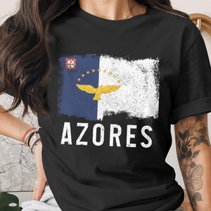 May include: Black t-shirt featuring a distressed design of the Azores flag. The flag is divided vertically, with a blue section on the left and a white section on the right. The word "AZORES" is printed in white below the flag.