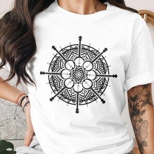 May include: White t-shirt featuring a black and white mandala design. The mandala is circular with a central starburst and banjo-like shapes radiating outwards. The shirt has short sleeves and a crew neck.