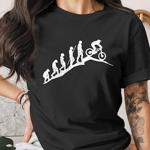 May include: Black t-shirt featuring a white graphic depicting the evolution of mountain biking. The design shows a progression from early hominids to a cyclist on a mountain bike. The shirt is a classic crew neck style.