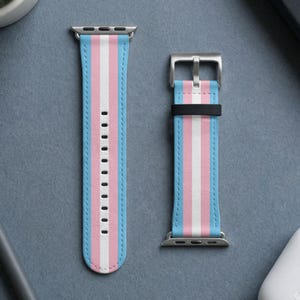 Puede incluir: Una correa de reloj de dos piezas con un patrón de rayas verticales en azul claro, rosa y blanco. La correa incluye conectores de metal plateado y una hebilla plateada con una trabilla negra, diseñada para un dispositivo smartwatch.