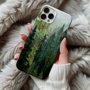 May include: A smartphone case featuring a forest scene with tall, green trees and a misty mountain backdrop. The case has a glossy finish and a protective design. The case is held in a person's hand.