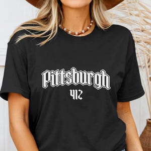 May include: Black t-shirt with the word "Pittsburgh" in a white, gothic-style font, and the number "412" below it. The shirt is a classic crew neck style.