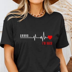 May include: Black t-shirt featuring a white heartbeat line design with the text "ANNNND..." and "I'M BACK" in red. The design includes a red heart at the end of the heartbeat line. The shirt is a classic crew neck style.