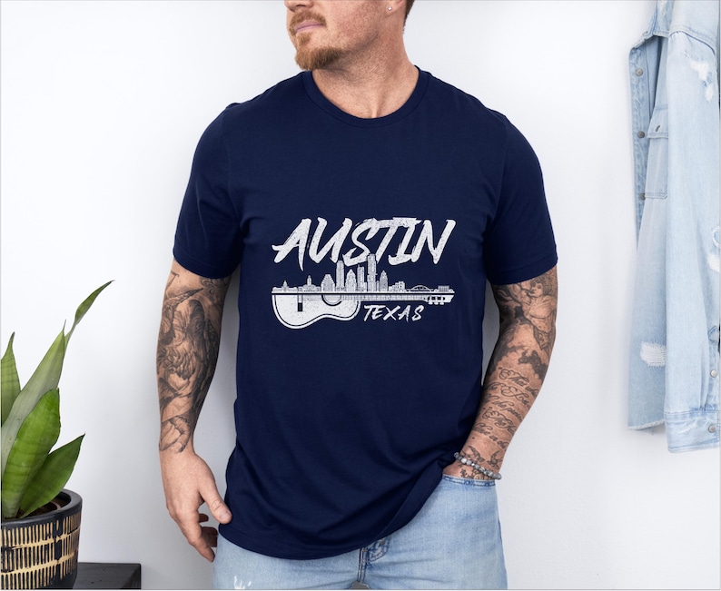 May include: A navy blue t-shirt with a white graphic of the Austin, Texas skyline with a guitar silhouette. The text "Austin Texas" is printed below the graphic.