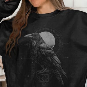 May include: A black long-sleeve sweatshirt featuring a detailed graphic print of a raven in profile. The bird is positioned in front of a circular moon shape and intricate geometric line work, all rendered in shades of gray and white.