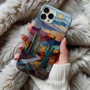 Puede incluir: Una funda de teléfono inteligente con un diseño de paisaje desértico. La funda tiene un estilo de vidriera con un cielo azul, un sol amarillo y cactus verdes. La paleta de colores incluye tonos de azul, morado, naranja y marrón. La funda se sostiene en una mano.