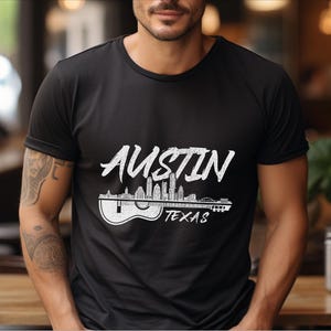May include: Black t-shirt with a white graphic of the Austin, Texas skyline with the word "Austin" in a stylized font and the words "Texas" below the skyline. The skyline is depicted as a guitar with the buildings as the fretboard.