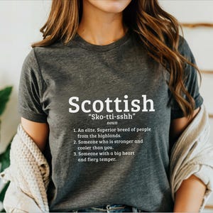 May include: A dark gray t-shirt with white text that reads "Scottish" and "Sko-tti-sshh" with a definition of the word. The definition includes the words "elite, superior breed of people, highlands, stronger, cooler, big heart, fiery temper."