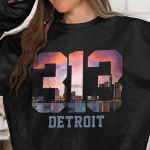 May include: Black sweatshirt featuring the number "313" in a stylized font with a sunset-sky background and a cityscape silhouette. The word "DETROIT" is printed below in a light blue font.