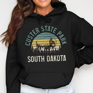 May include: A black hooded sweatshirt featuring a graphic print of a bison silhouette against a sunset background with pine trees. The text reads CUSTER STATE PARK and SOUTH DAKOTA in a distressed font. The hoodie includes a front kangaroo pocket.