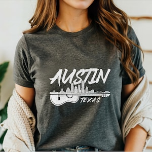 May include: A dark gray t-shirt with a white graphic design of the Austin, Texas skyline with a guitar silhouette. The text "Austin, Texas" is printed on the shirt.