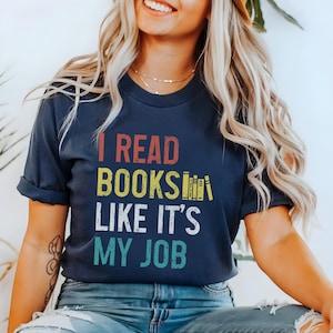 May include: A navy blue t-shirt with the text "I read books like it's my job" in a vintage style font. The text is in yellow, red, and blue. There are stacked books in the text.