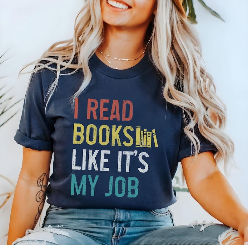 May include: A navy blue t-shirt with the text "I read books like it's my job" in a vintage style font. The text is in yellow, red, and blue. There are stacked books in the text.