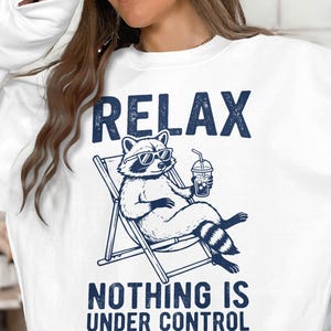 May include: A white long-sleeve sweatshirt featuring a dark blue graphic of a raccoon wearing sunglasses, sitting in a lounge chair and holding a drink. The text above reads RELAX and the text below reads NOTHING IS UNDER CONTROL.