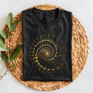 May include: Black t-shirt featuring a golden spiral design. The spiral is intricate, with detailed lines and dots, creating a mesmerizing pattern. The shirt is folded on a woven, circular mat with a sprig of green leaves.