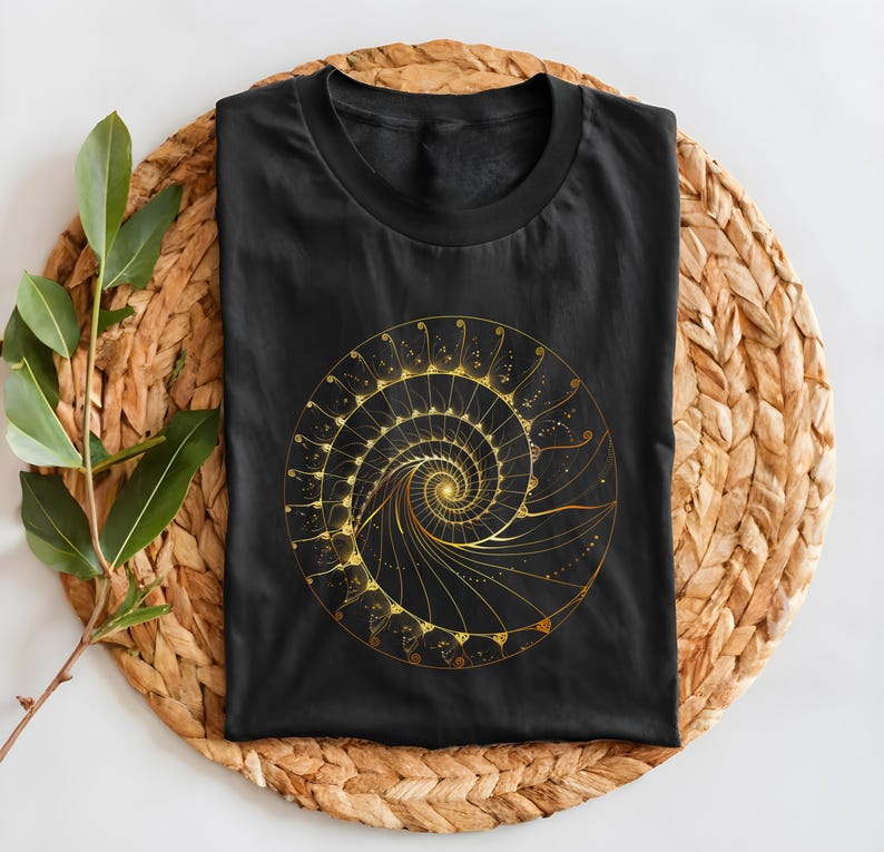 May include: Black t-shirt featuring a golden spiral design. The spiral is intricate, with detailed lines and dots, creating a mesmerizing pattern. The shirt is folded on a woven, circular mat with a sprig of green leaves.