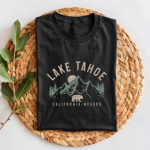 Puede incluir: Camiseta negra con un gr&aacute;fico de estilo vintage de monta&ntilde;as, un oso y un sol. El texto "LAKE TAHOE CALIFORNIA-NEVADA" est&aacute; impreso en la camiseta.