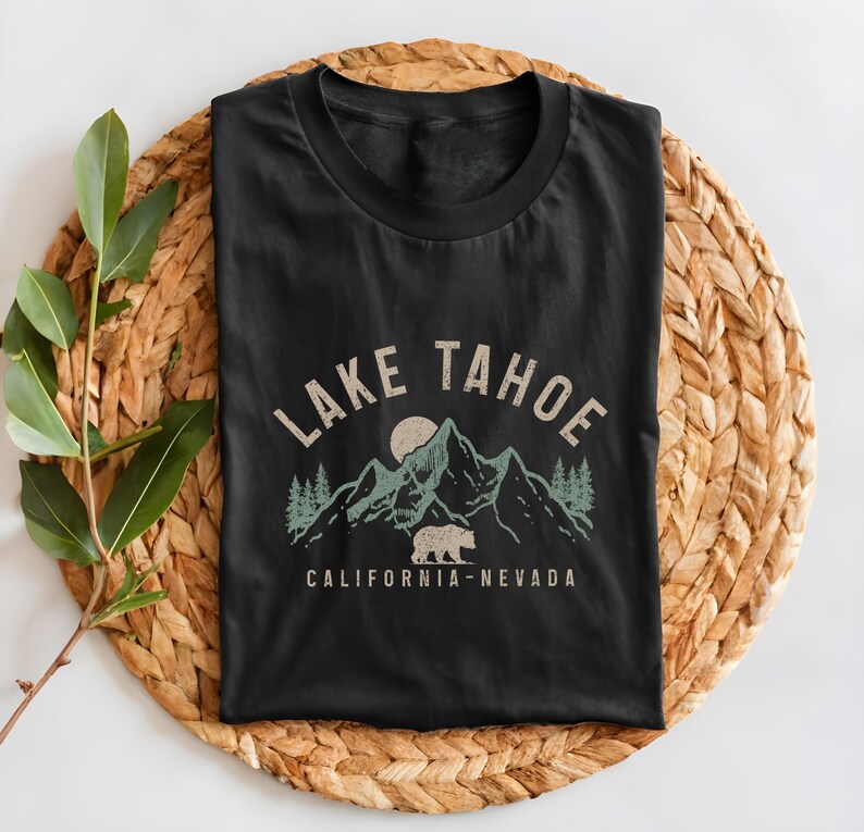 Puede incluir: Camiseta negra con un gr&aacute;fico de estilo vintage de monta&ntilde;as, un oso y un sol. El texto "LAKE TAHOE CALIFORNIA-NEVADA" est&aacute; impreso en la camiseta.