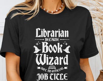 May include: Black t-shirt with white text that reads "Librarian because Book Wizard isn't an official job title." The design includes witch hats and stars. The shirt is a crew neck.