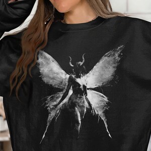 May include: A black long-sleeve sweatshirt featuring a large, white, distressed-style graphic print on the front. The graphic depicts a central figure with large, detailed wings and small horns, rendered in a monochromatic, sketch-like aesthetic.