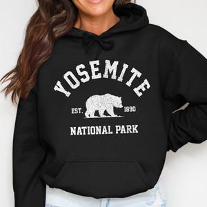 Op de afbeelding: Zwarte hoodie met een witte print met de tekst "YOSEMITE NATIONAL PARK" en een berensilhouet. De tekst "EST. 1890" is ook inbegrepen. De hoodie heeft een voorzak.
