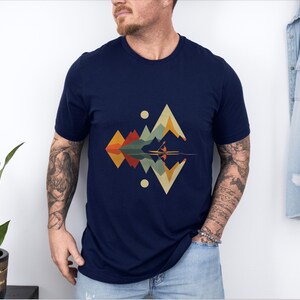 May include: A navy blue t-shirt with a geometric design featuring a mountain range and a reflection in water. The design is in shades of green, brown, orange, and yellow.