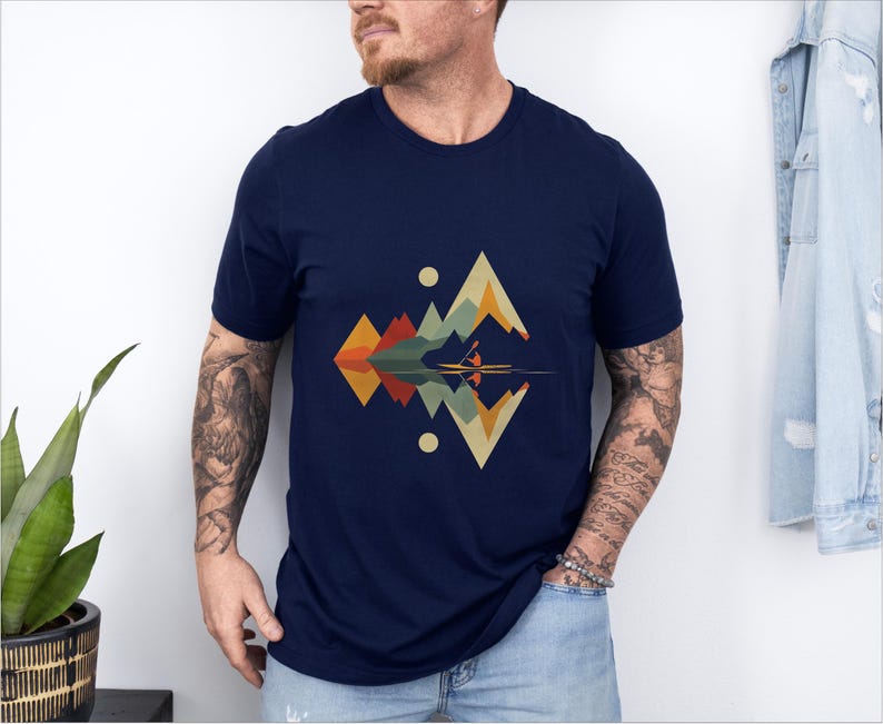 May include: A navy blue t-shirt with a geometric design featuring a mountain range and a reflection in water. The design is in shades of green, brown, orange, and yellow.