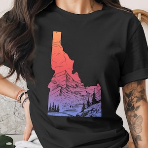 May include: Black t-shirt featuring a colorful graphic of the state of Idaho. The design showcases a mountain landscape with a gradient of orange, pink, and purple hues. The shirt is a classic crew neck style.