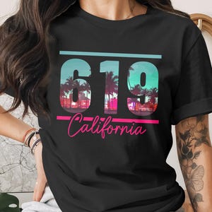 以下が含まれることがあります： 黒いTシャツ。数字の「619」は、ティールとピンクのグラデーションで、ヤシの木のシルエットのデザイン。 下にはピンクの筆記体で「California」の文字。カジュアルウェアです。