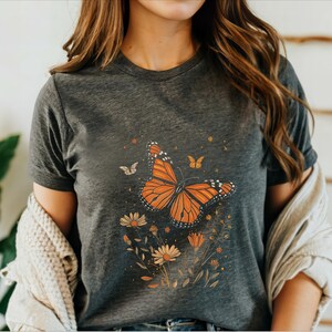 K&ouml;nnte beinhalten: Ein dunkelgraues T-Shirt mit einem Aufdruck eines orange-schwarzen Schmetterlings, umgeben von gelben und braunen Blumen.