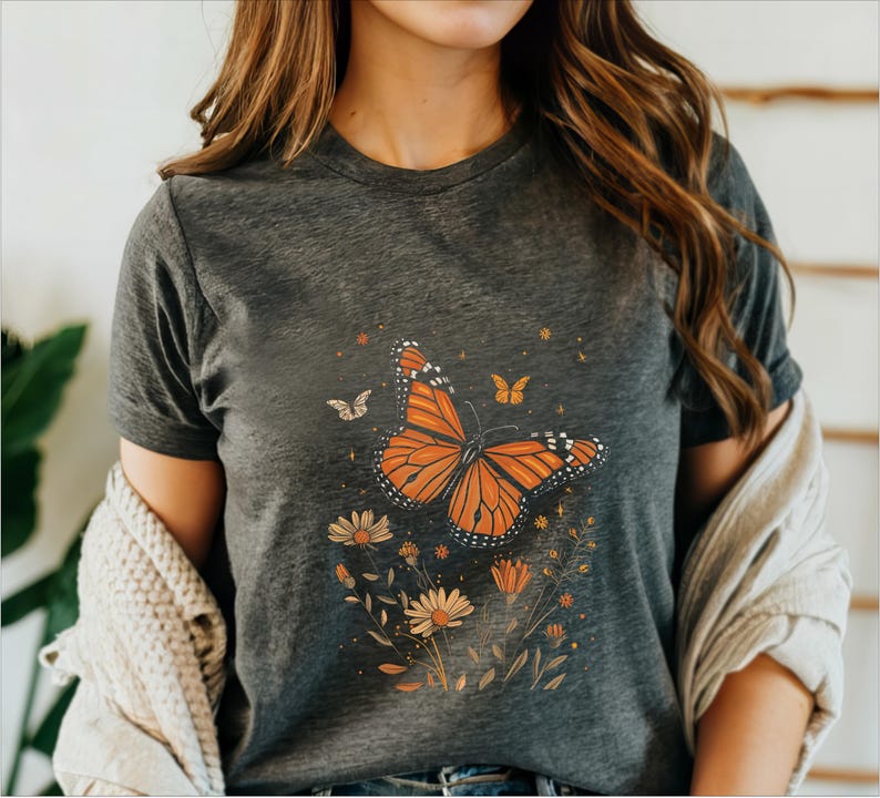 K&ouml;nnte beinhalten: Ein dunkelgraues T-Shirt mit einem Aufdruck eines orange-schwarzen Schmetterlings, umgeben von gelben und braunen Blumen.