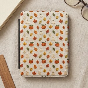 May include: A rectangular e-reader case featuring a cream-colored fabric cover with an embroidered pattern of small orange pumpkins, various autumn leaves in shades of red, yellow, and green, and tiny white ghosts scattered across the surface.