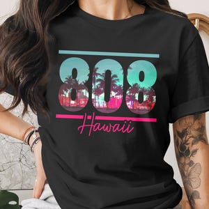 May include: Black t-shirt featuring a colorful graphic design. The design includes the numbers "808" in a teal and blue gradient, with a tropical scene inside each number. Below the numbers, the word "Hawaii" is written in pink script.
