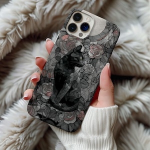 Puede incluir: Una funda de teléfono con un diseño de gato negro. La funda tiene un patrón floral gris y negro con un efecto de vidriera. El gato está sentado y mirando hacia un lado. La funda la sostiene una persona que lleva un jersey blanco.