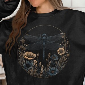 May include: Black sweatshirt featuring a circular design with a dragonfly and floral elements. The dragonfly is dark blue, and the flowers are in shades of blue and beige. The design is framed by a gold-colored circle.